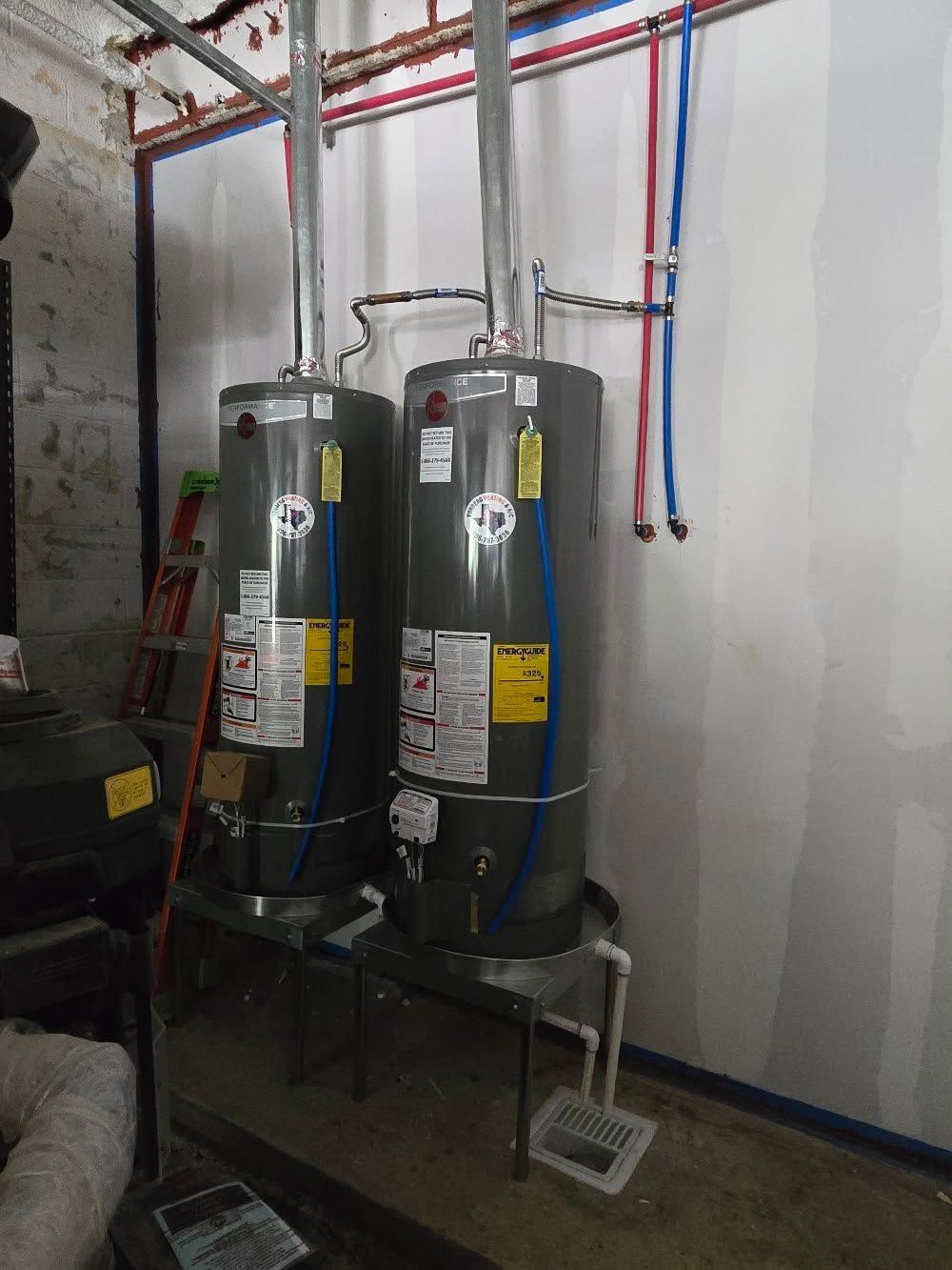 Two gray water heaters with colored pipes in a utility room against a white wall
