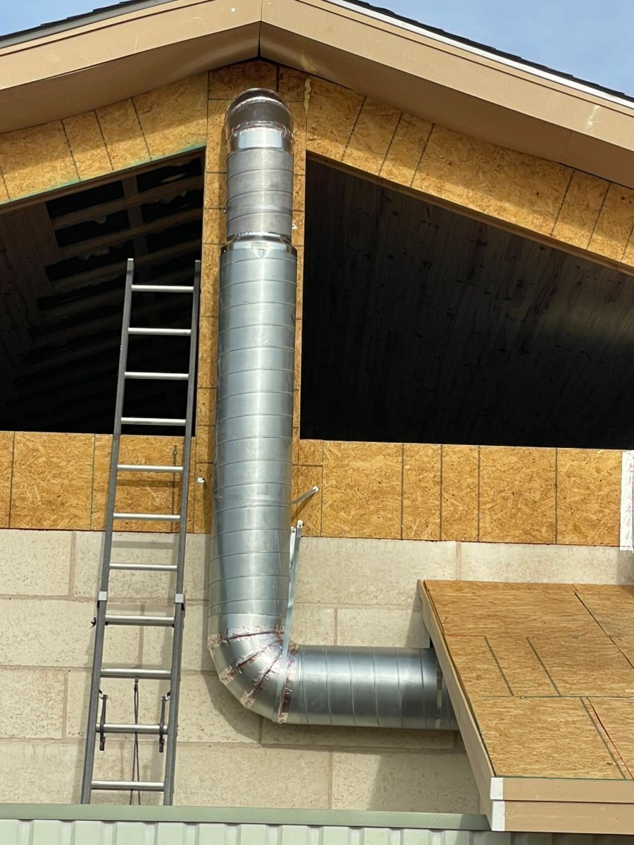 Silver metal vent pipe on a building exterior with a ladder leaning beside it and boarded roof opening
