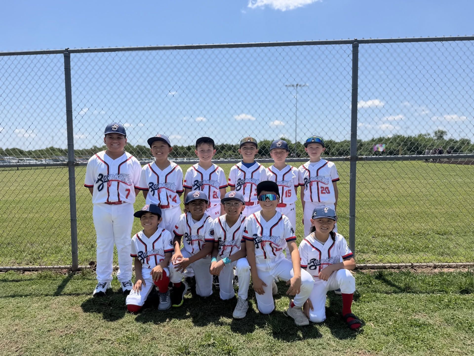 Youth baseball team posing on a field in white uniforms with a chain-link fence behind them