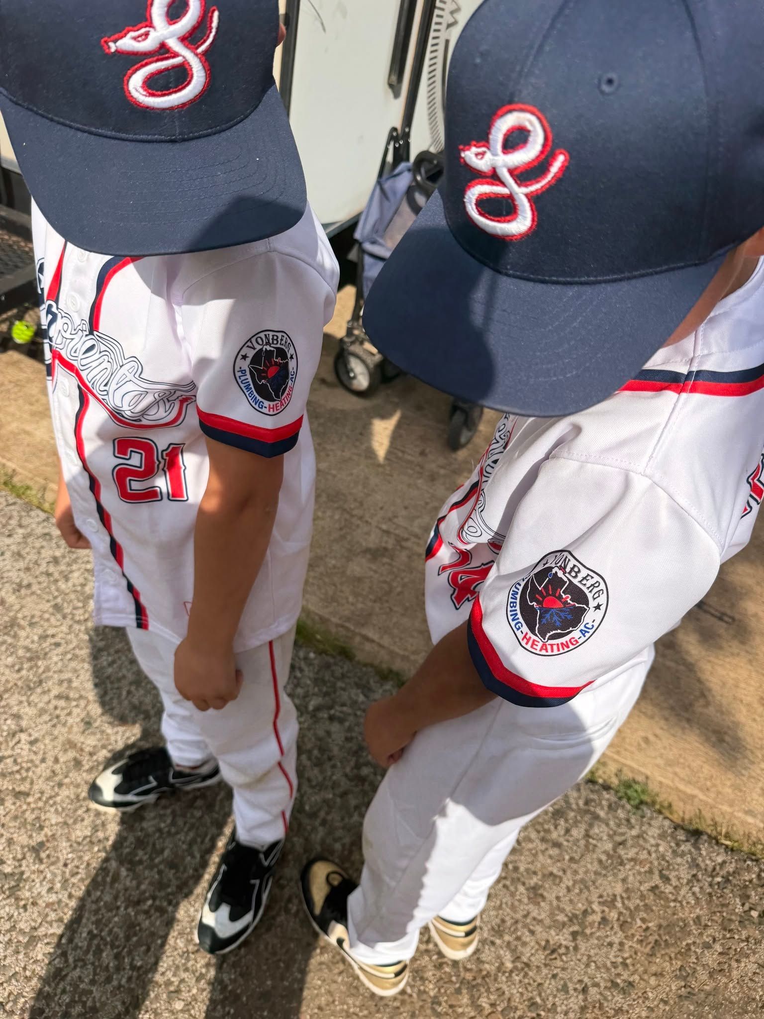 Two youth baseball players in white and navy uniforms standing on a field, viewed from above.