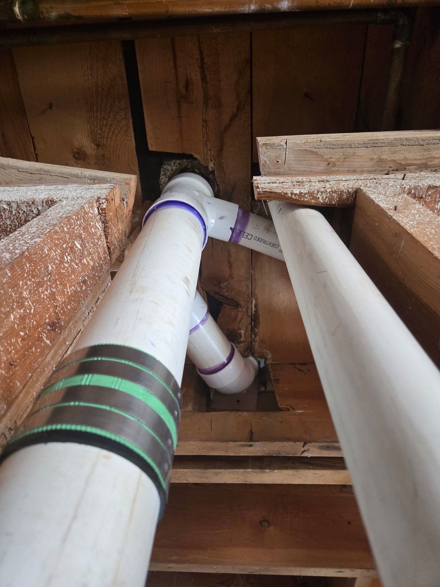 White PVC pipes and purple-joint plumbing running through exposed wooden floor joists.