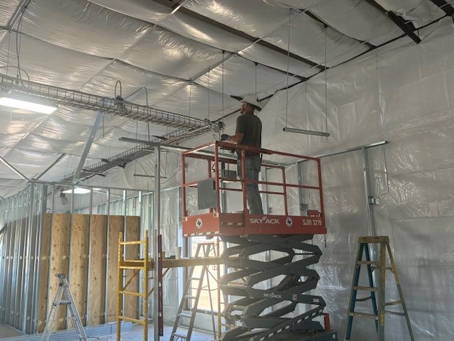 Worker on scissor lift installing insulation in a framed room with plastic-covered walls and ceiling