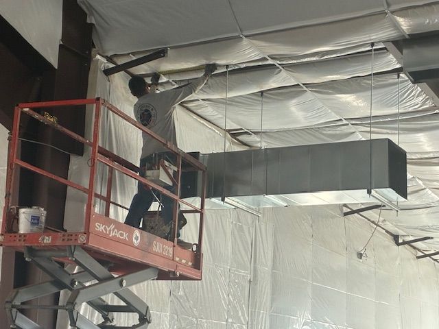 Worker on a red lift installing a large rectangular HVAC duct near a ceiling.