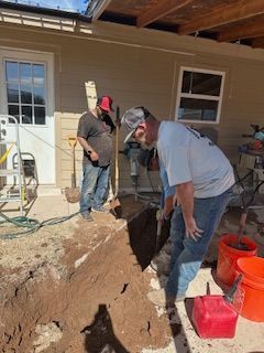 Two workers dig a trench beside a tan house, with tools and orange buckets nearby.