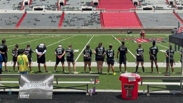 Football players standing on the sideline in black uniforms, facing the field in an empty stadium