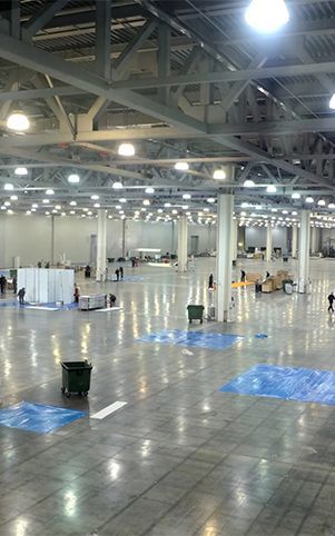 Large, empty exhibition hall with many lights and tall support beams. Workers are visible, and blue tarp patches dot the floor.