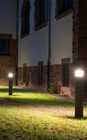 Two outdoor path lights illuminating a grassy area with a stone building in the background at night.