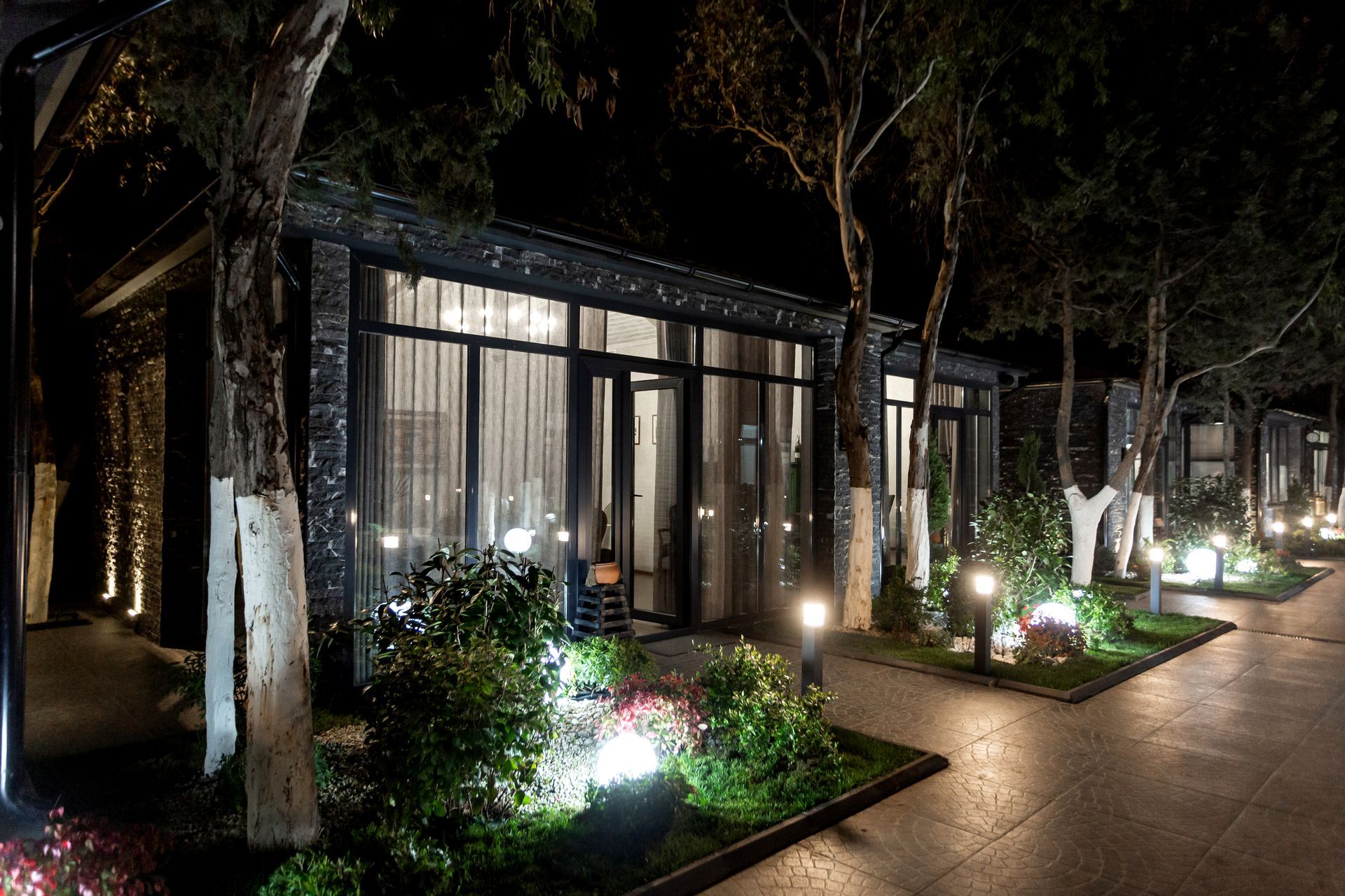 Night view of lit outdoor restaurant buildings, trees lining the walkway, garden lights.