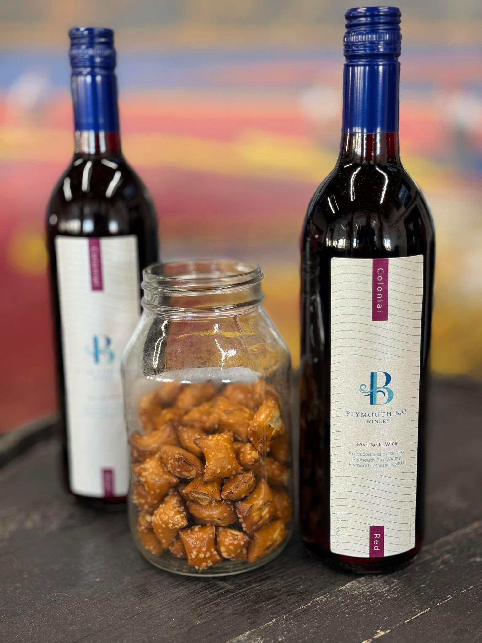 Two wine bottles with blue caps and a jar of pretzels on a table.