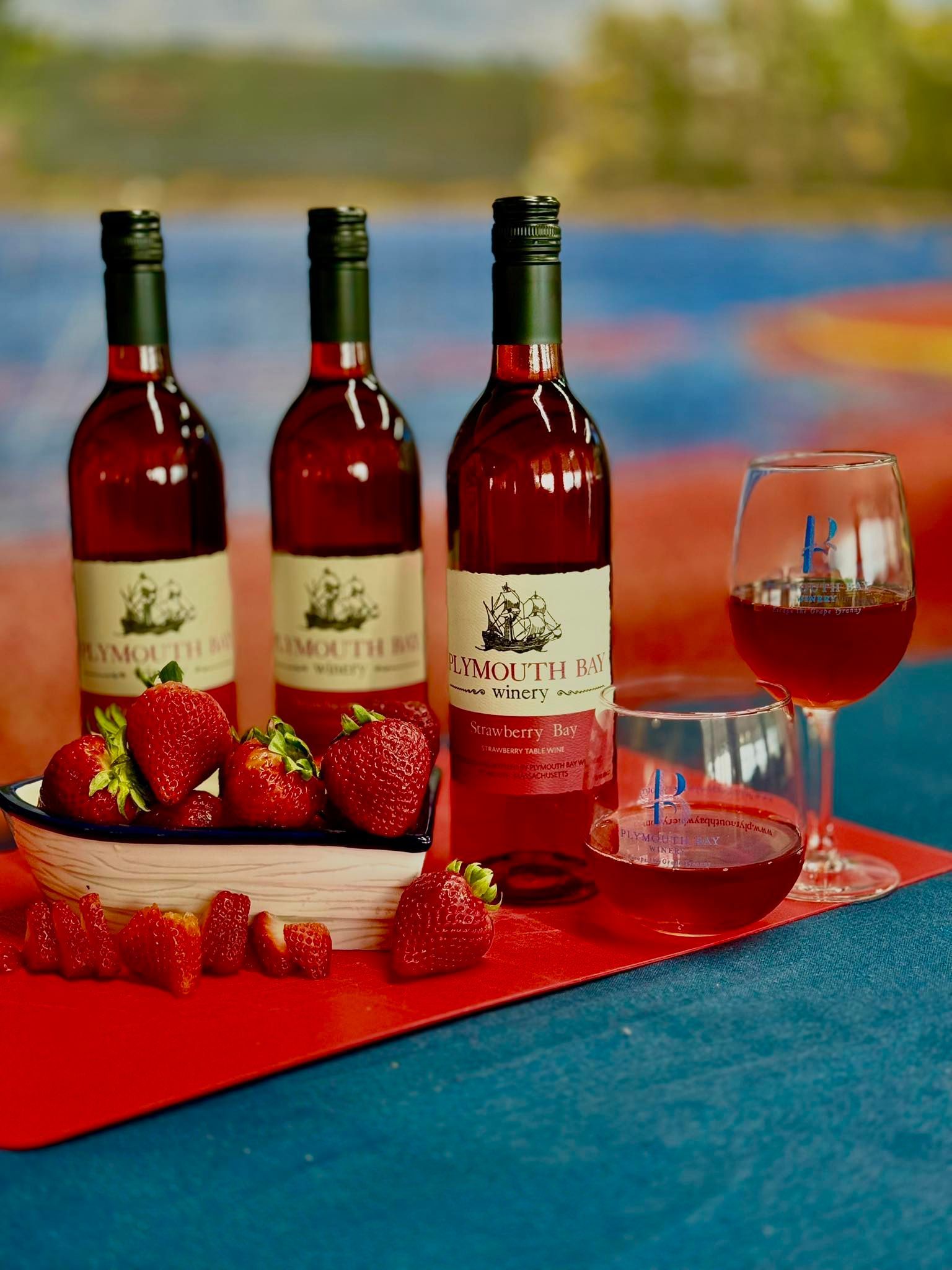 Three bottles of red wine and a glass, strawberries on a red surface, blue background.
