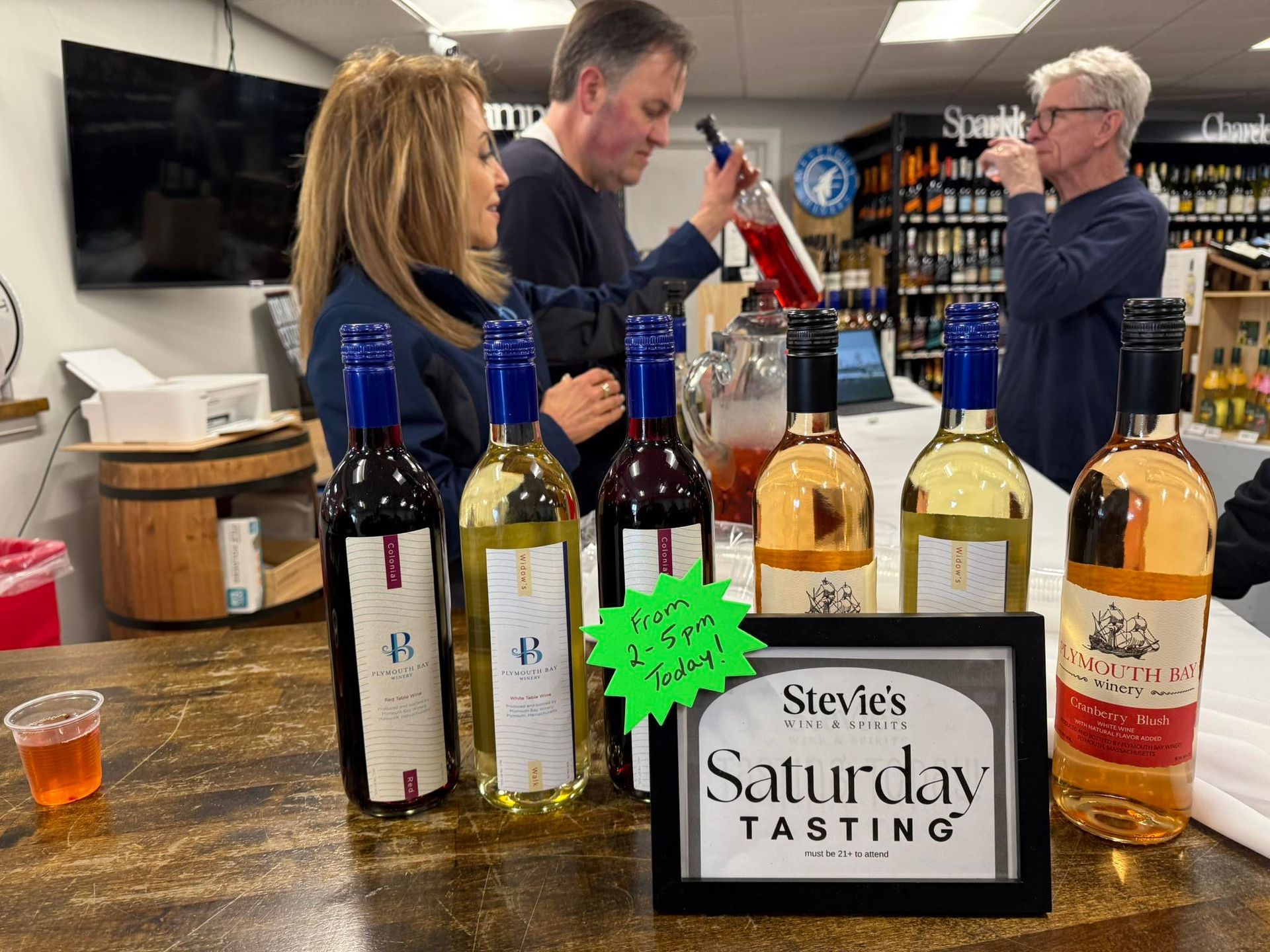 Wine tasting event at Stevie's, bottles on a bar. People sample wine. Sign: Saturday Tasting.