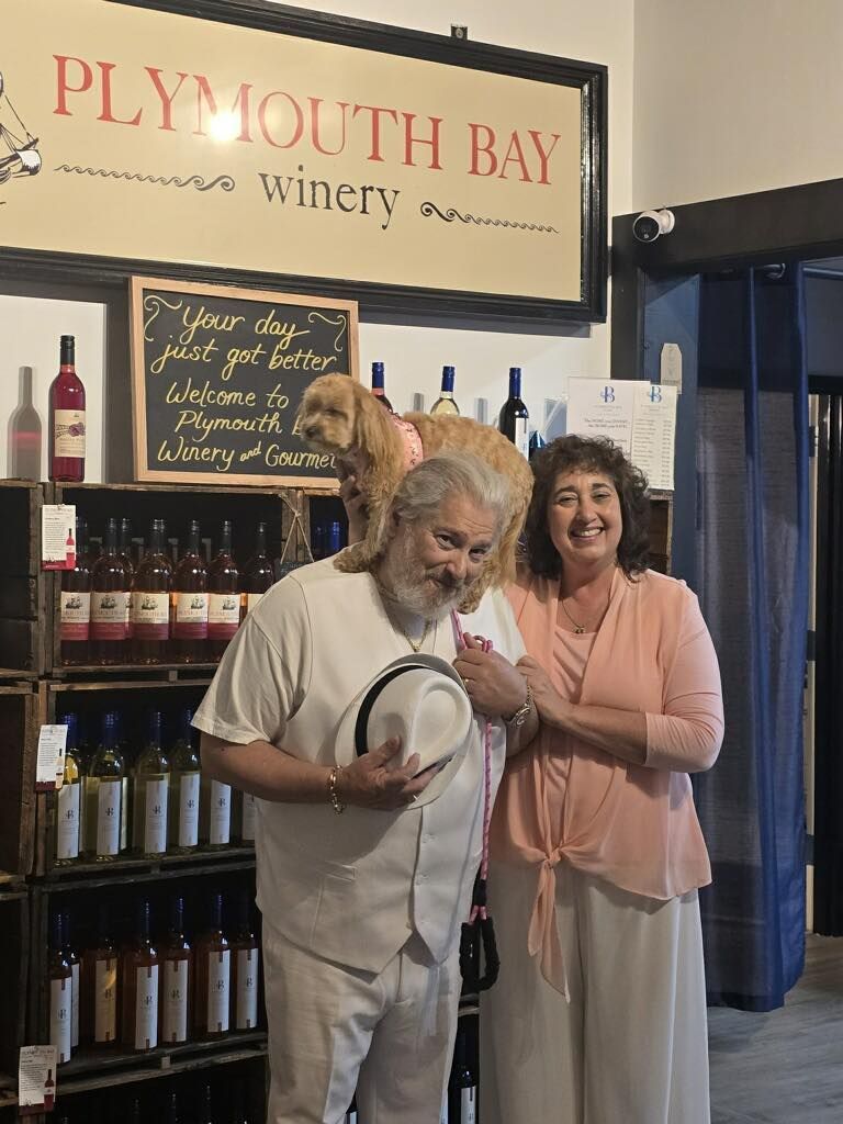 Man and woman pose at Plymouth Bay Winery with dog on man's shoulders, surrounded by wine bottles.