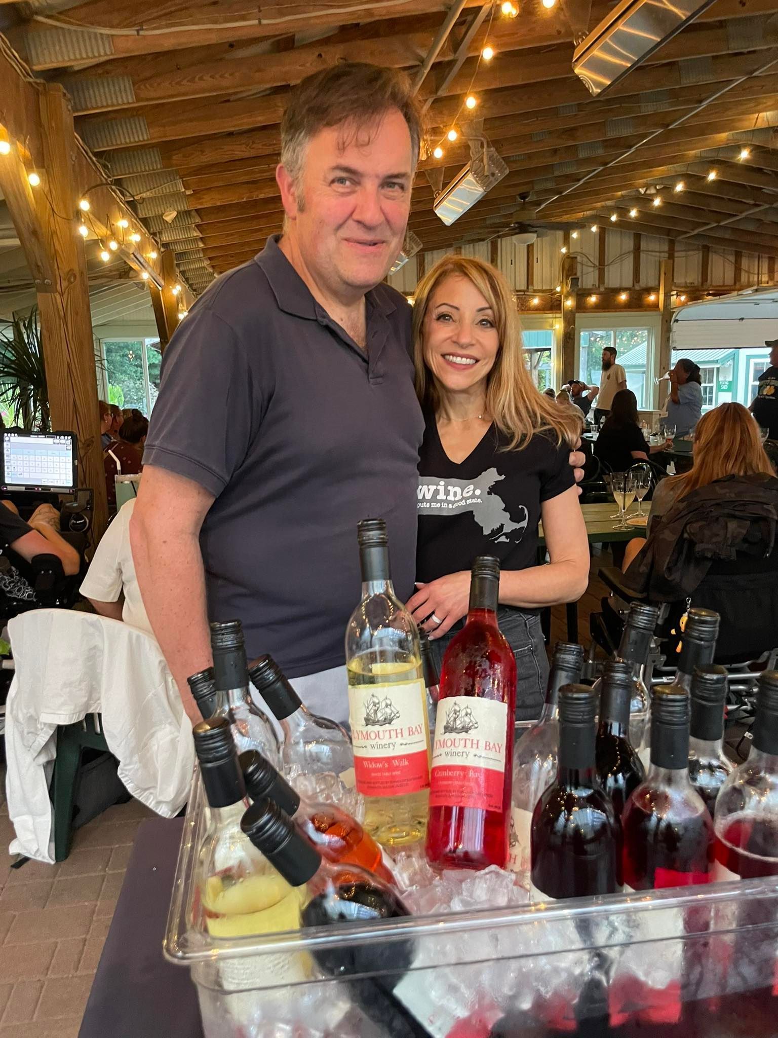 Man and woman stand behind a table with wine bottles, smiling. Lit outdoor setting.