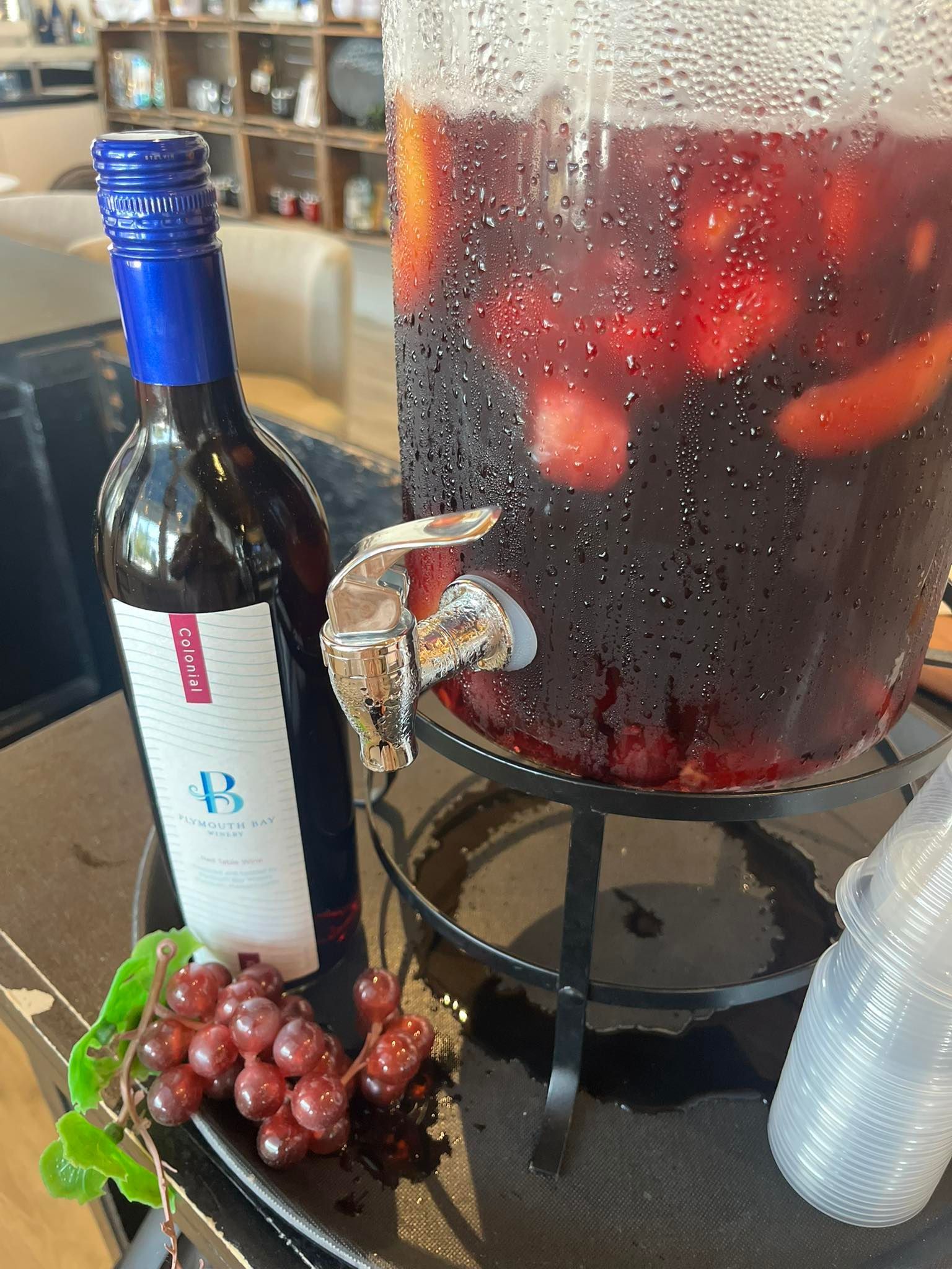 Red wine bottle and a sangria dispenser with grapes on a table.