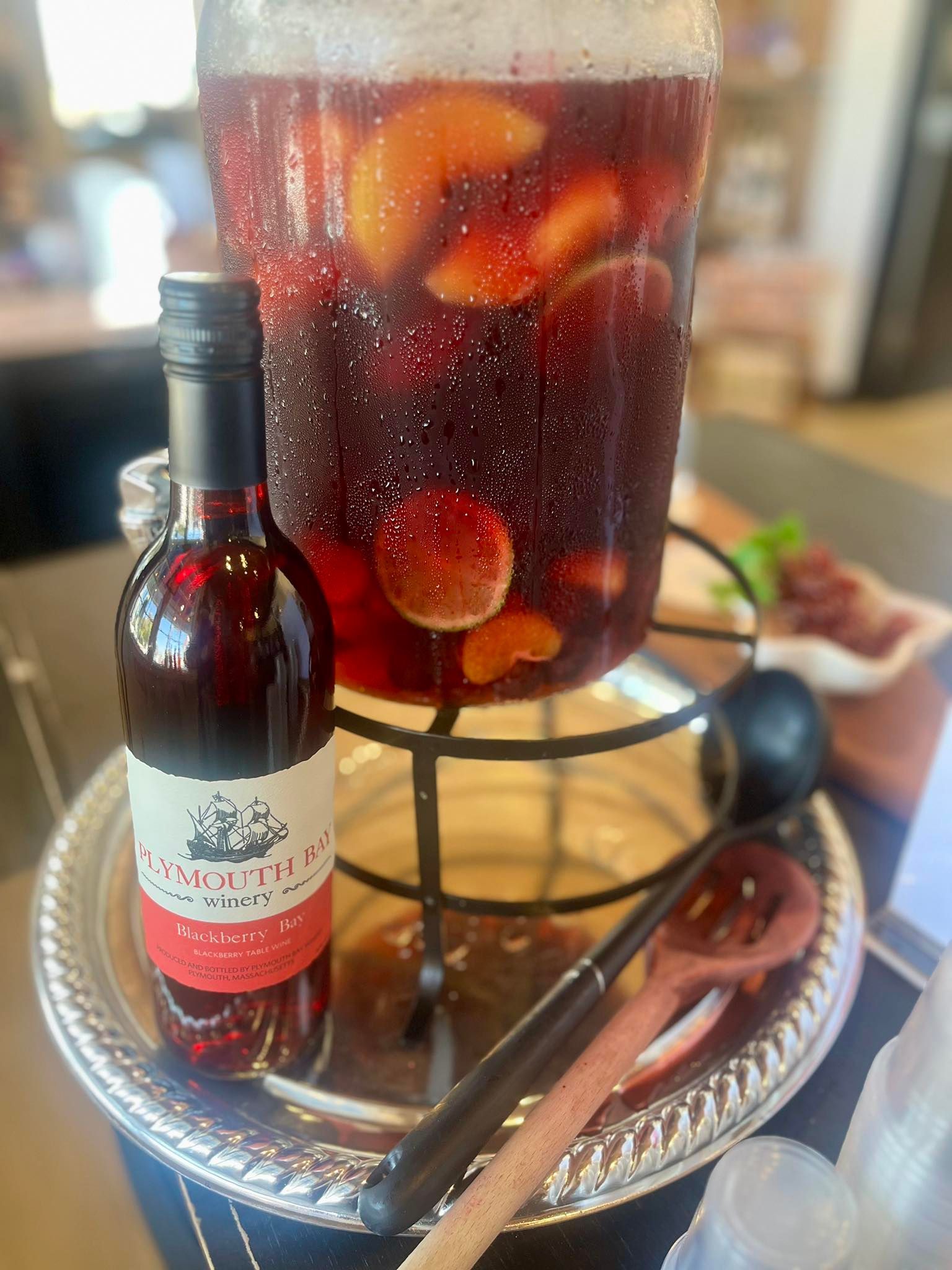 Sangria dispenser with fruit, a bottle of red wine, and a serving spoon on a silver platter.