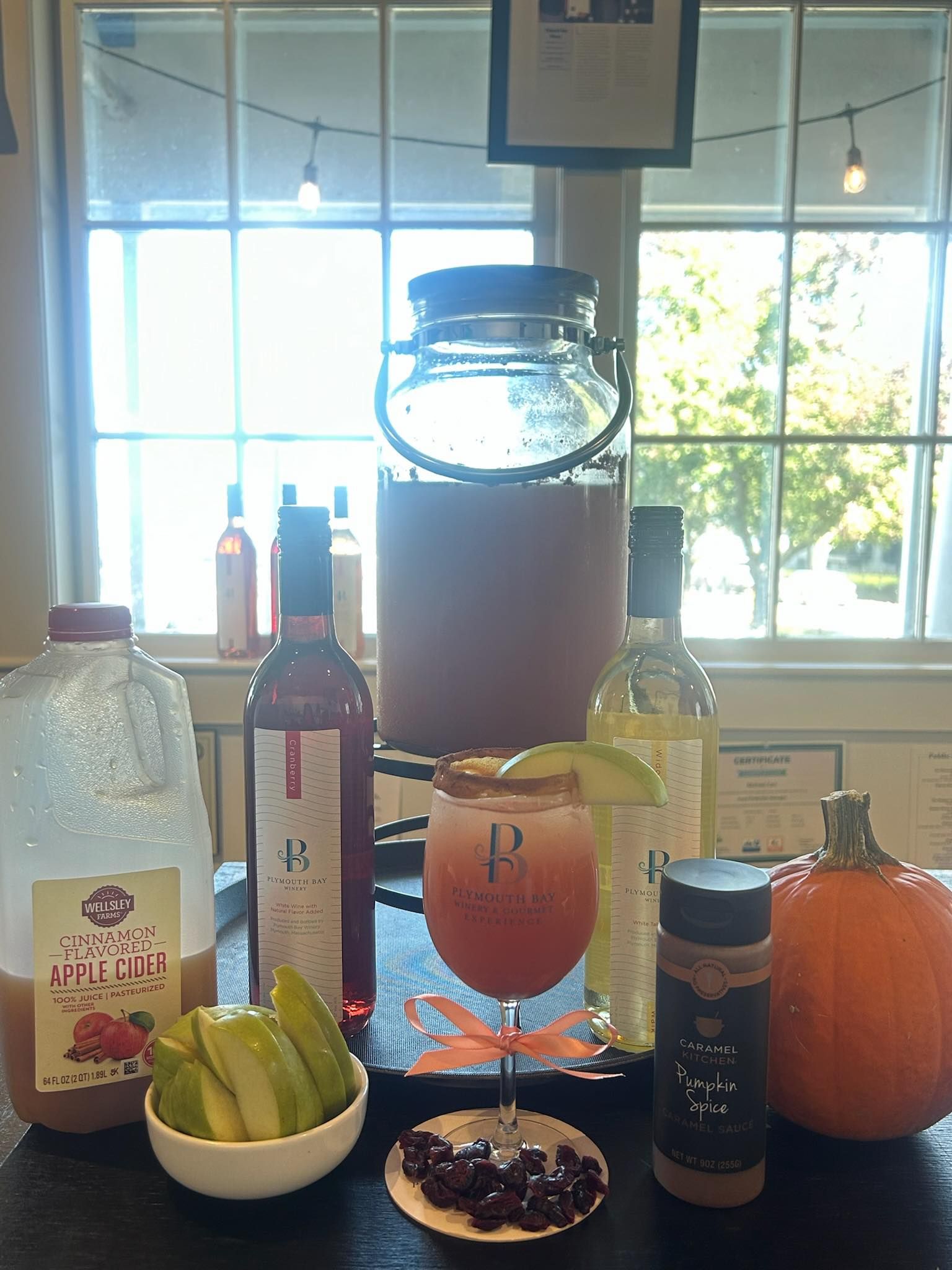 Fall drink setup with cider, wine, cocktail, pumpkin, spices, and fruit on a table by a window.