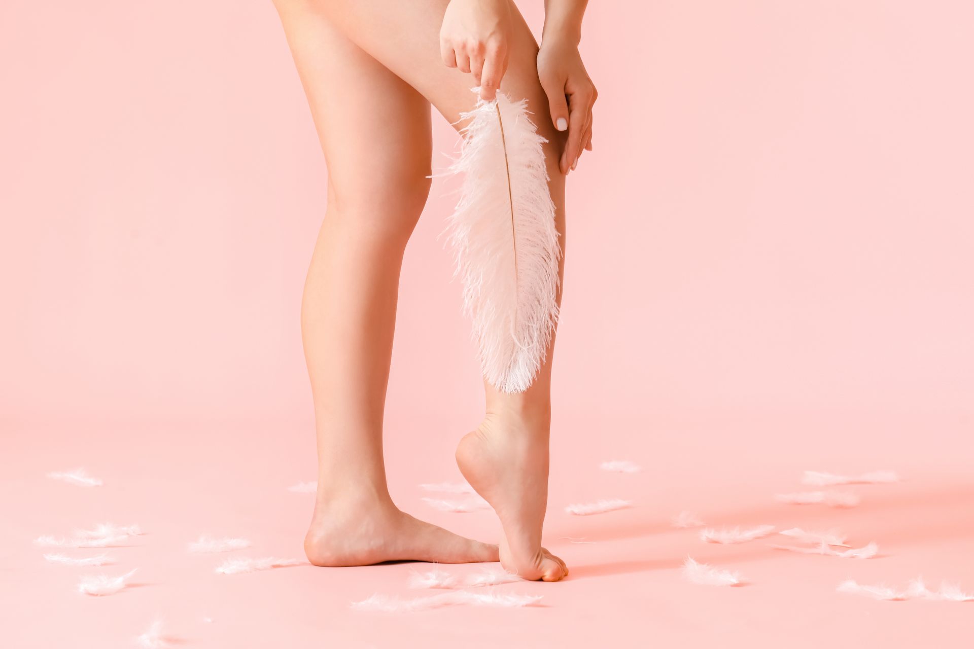 A woman is holding a feather on her leg on a pink background.