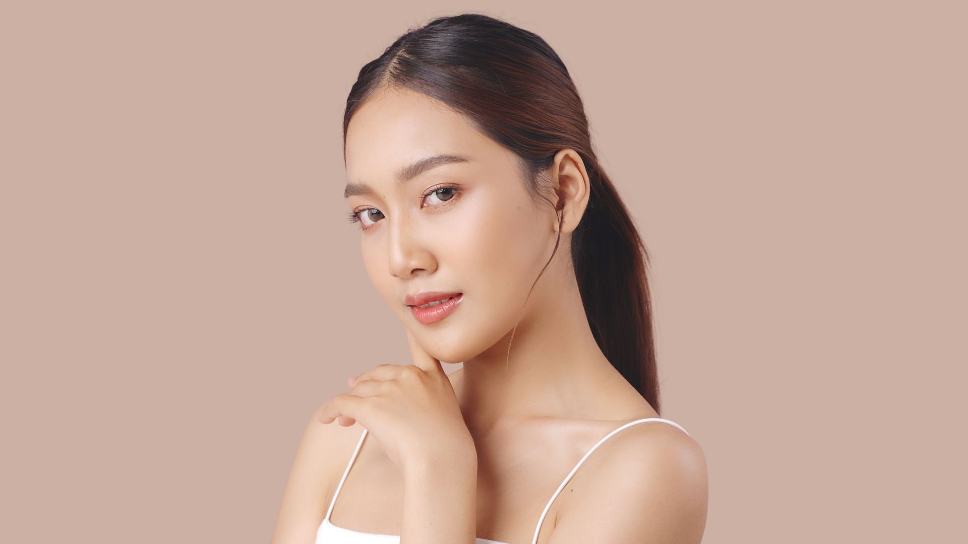 A woman with a ponytail is touching her face and looking at the camera.