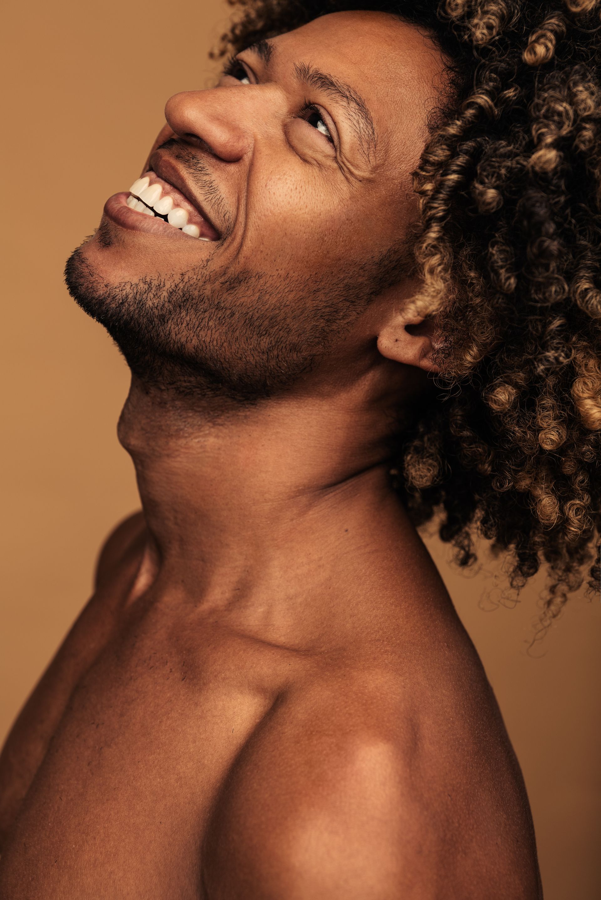 A shirtless man with curly hair is smiling and looking up.