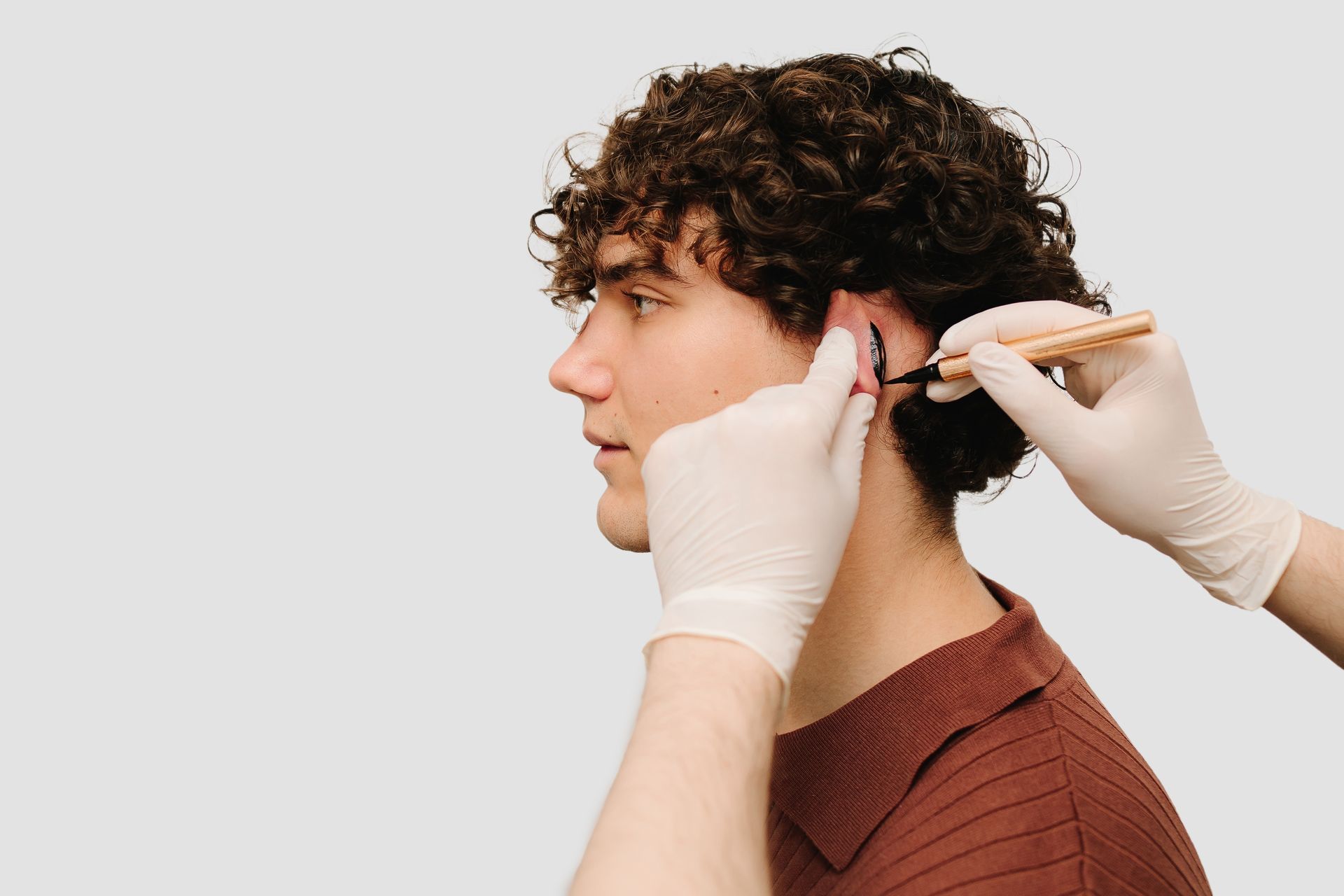 A man with curly hair is getting his ear pierced by a person wearing gloves.