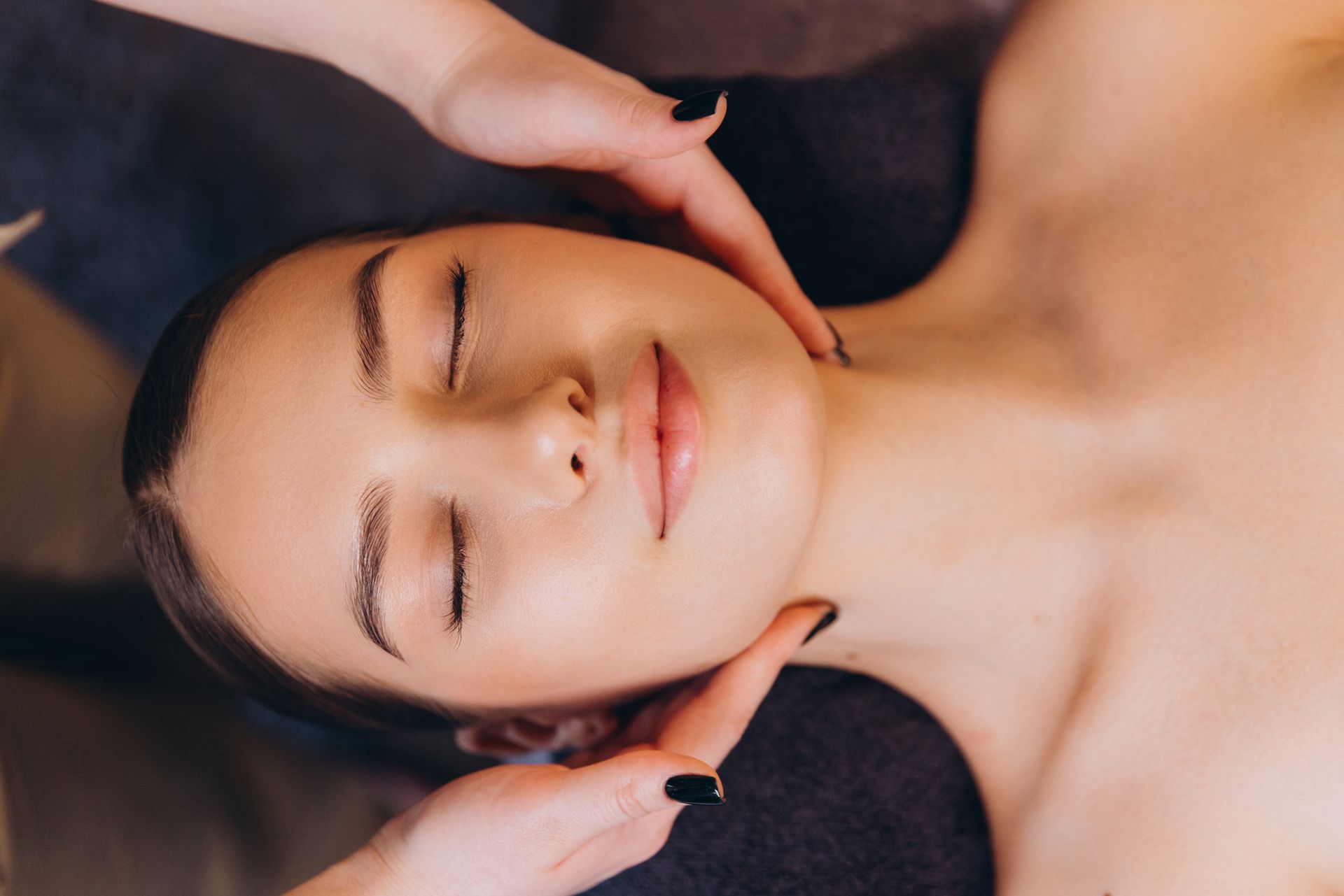 Woman receiving a facial massage with eyes closed, surrounded by hands on her face.