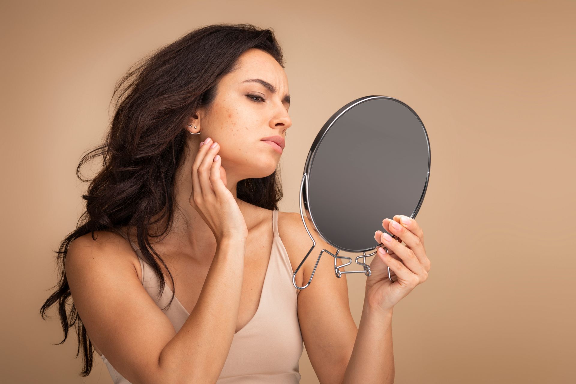 Woman applying makeup while looking into a handheld mirror against a beige background