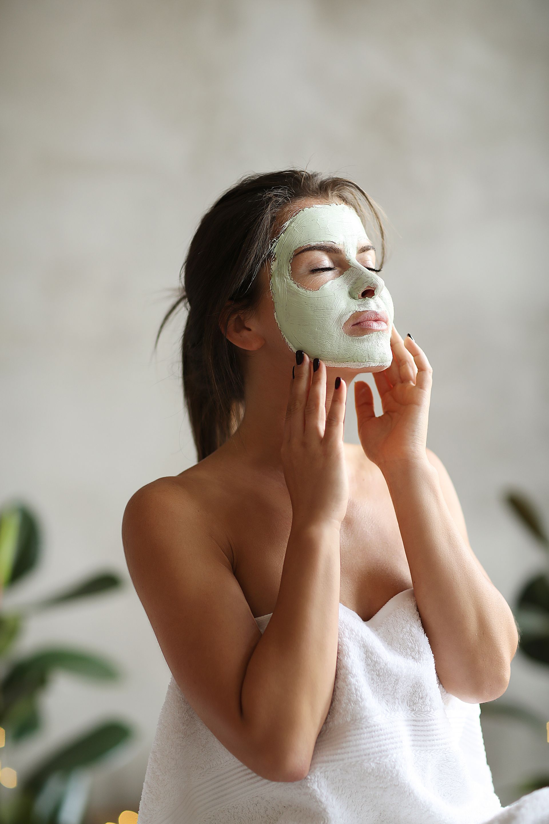 A woman wrapped in a towel is applying a clay mask to her face.