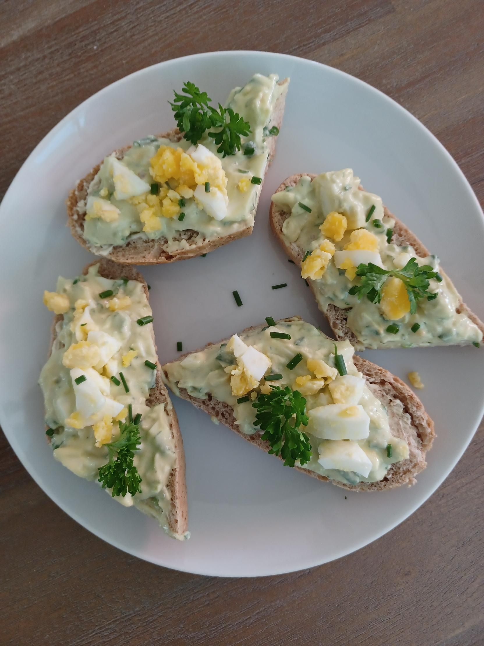 Scroll verder voor het recept Eiersalade op sneetjes brood, gegarneerd met peterselie en bieslook, op een wit bord.