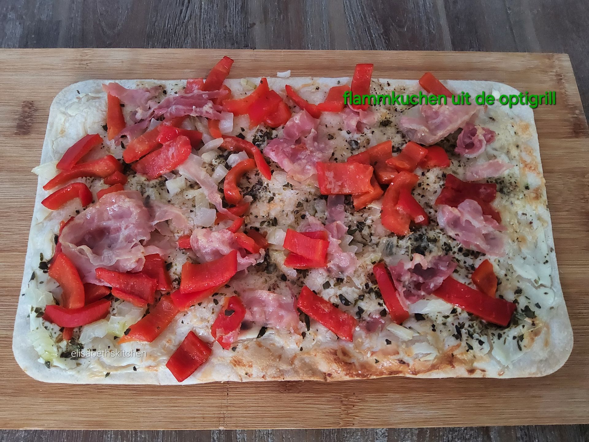 Scroll verder voor het recept Flammkuchen met rode paprika, spek en kruiden op een houten plank.