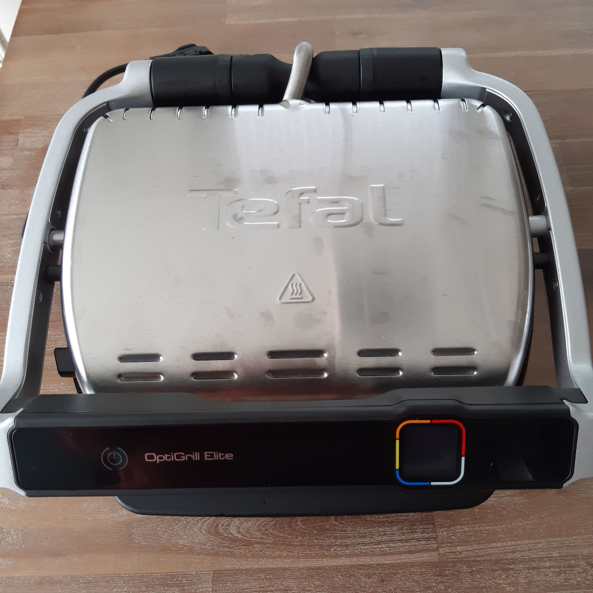 Scroll door voor het recept Tefal OptiGrill Elite op een houten ondergrond. Gesloten, zilverkleurige grill met zwarte voet, handgreep en bedieningspaneel.