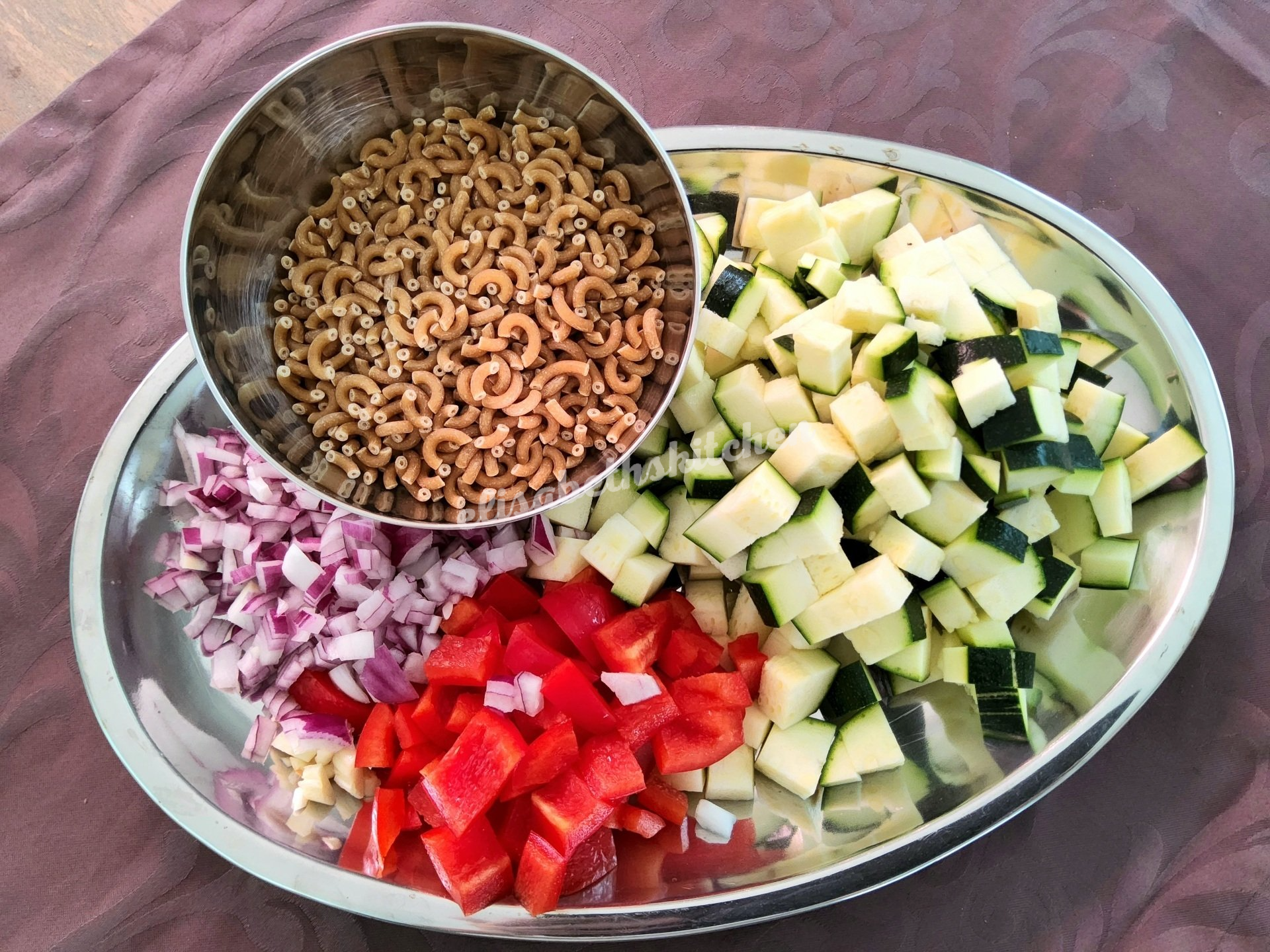 Ingrediënten voor een gerecht: pasta, in blokjes gesneden courgette, rode paprika, rode ui, in een metalen kom en op een schaal.