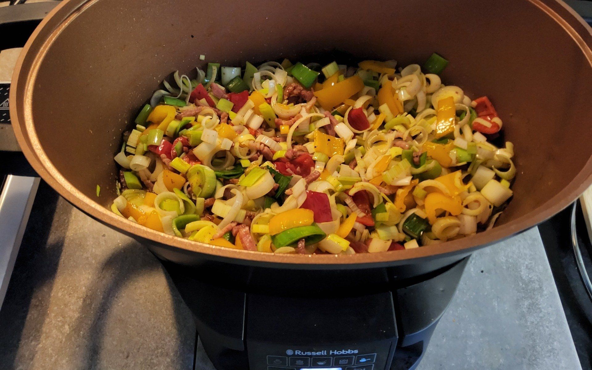 Groenten (prei, paprika's, tomaten) worden gekookt in een koperkleurige pan op het fornuis.