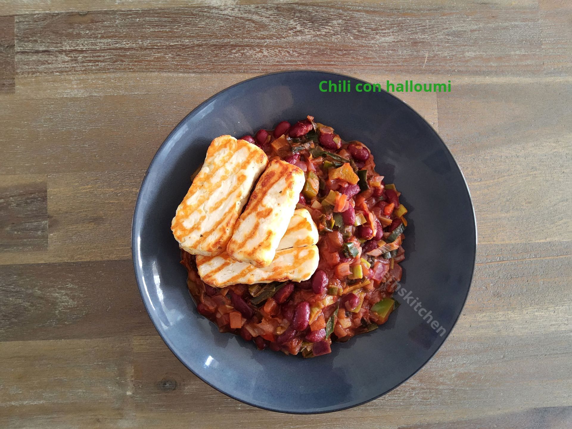 Scroll door voor het recept Chili con halloumi op een donker bord, met rode kidneybonen, groenten en gegrilde kaas.