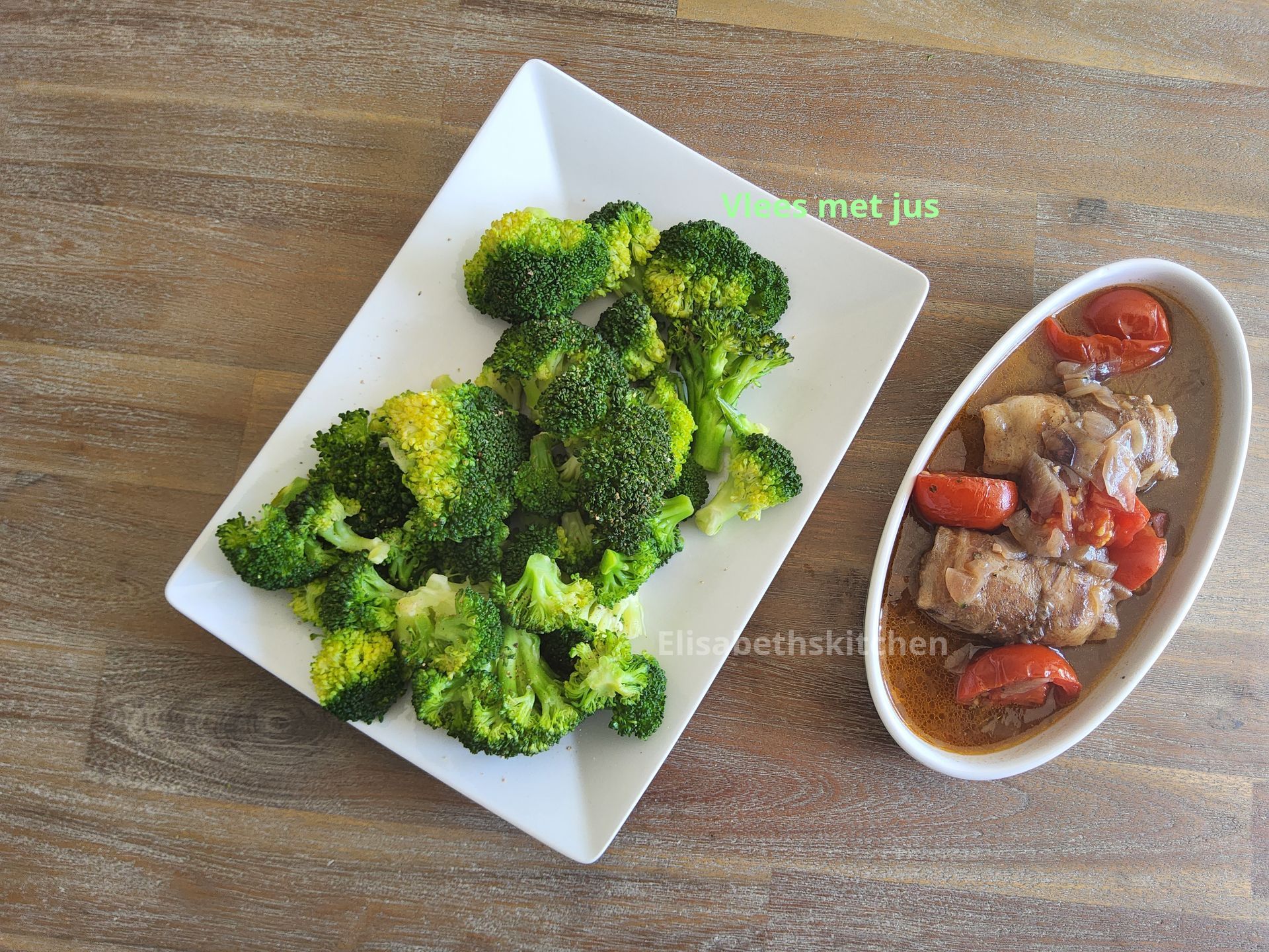 Broccoliroosjes op een wit bord, naast een schaal met vlees en tomaten in saus. Houten tafel.