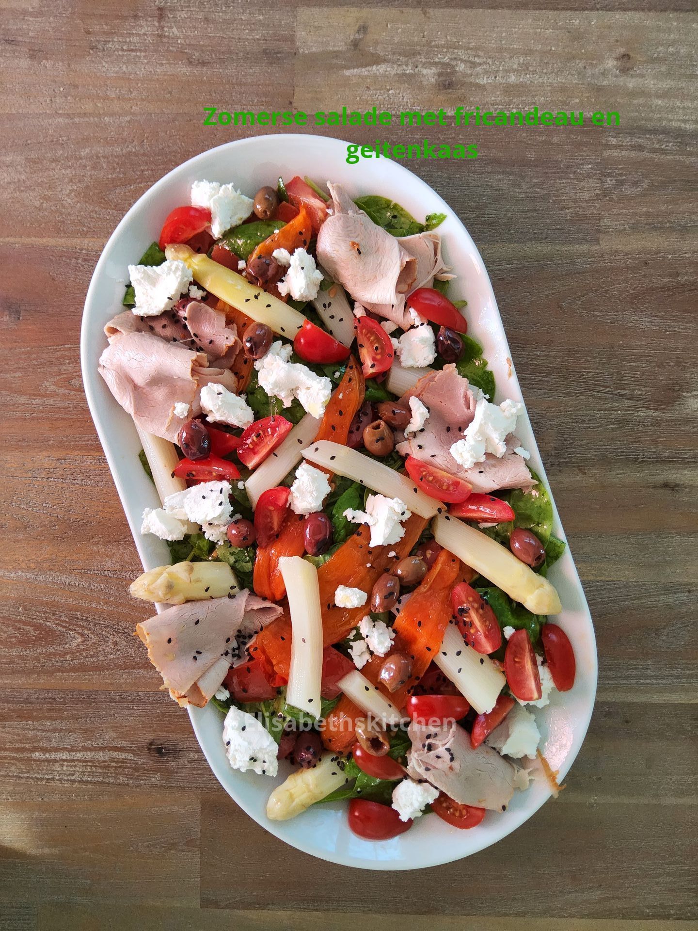Salade met asperges, tomaten, kaas, olijven en vlees op een wit ovaal bord, op een houten tafel.