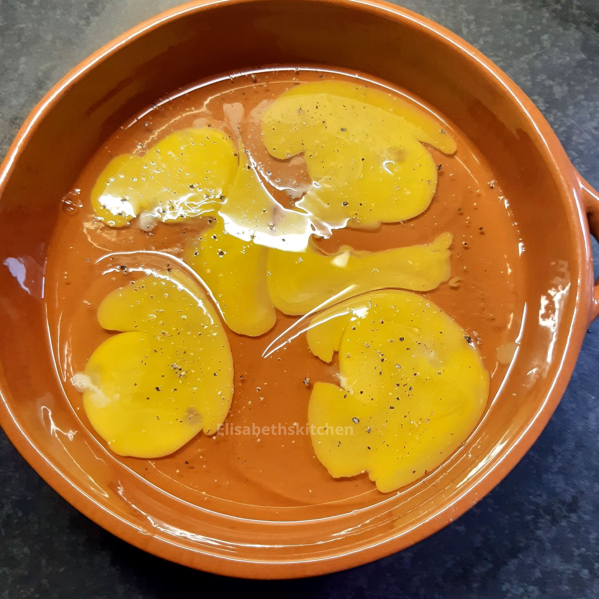 Gesneden aardappelen in een bruine ovenschaal met olie, gekruid met zwarte peper.