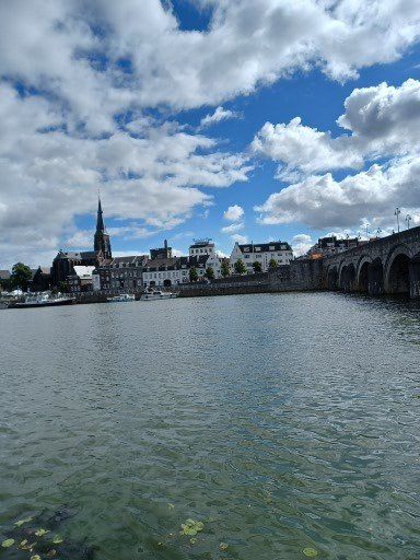 Stadsgezicht met water op de voorgrond en een bewolkte hemel. Brug, gebouwen en een kerktoren zijn zichtbaar.