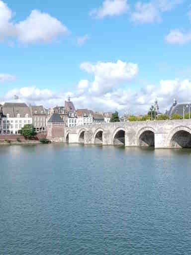 Een stenen brug met boogvormige openingen overspant een brede, blauwe rivier; aan de overkant staan gebouwen onder een zonnige hemel.