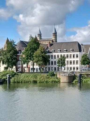 Gebouwen en kerktorens aan een rivier, onder een blauwe hemel met wolken.