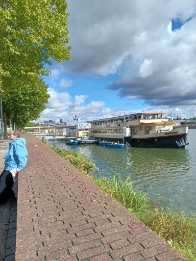 Een bakstenen pad langs een rivier met een aangemeerde boot, een blauwe lucht en een persoon in een blauwe jas.