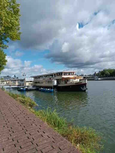 Een grote boot ligt aangemeerd aan een rivier naast een stenen wandelpad op een zonnige dag met wat bewolking.