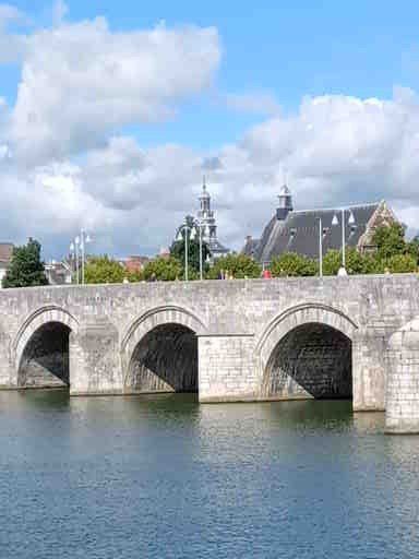 Stenen brug over het water, met boogvormige openingen. Gebouwen en een kerktoren zijn op de achtergrond te zien. Blauwe lucht met wolken.