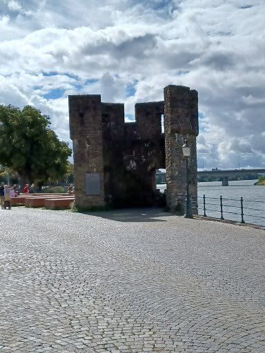 Een vervallen stenen toren op een geplaveid pad nabij een rivier, met in de verte een brug onder een bewolkte hemel.