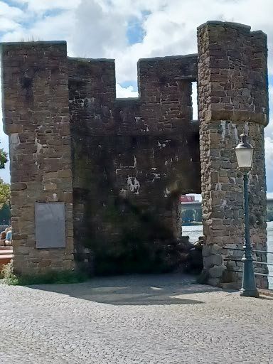 Ruïne van een stenen muur met een lantaarnpaal, tegen een blauwe hemel, aan de oever van een rivier.