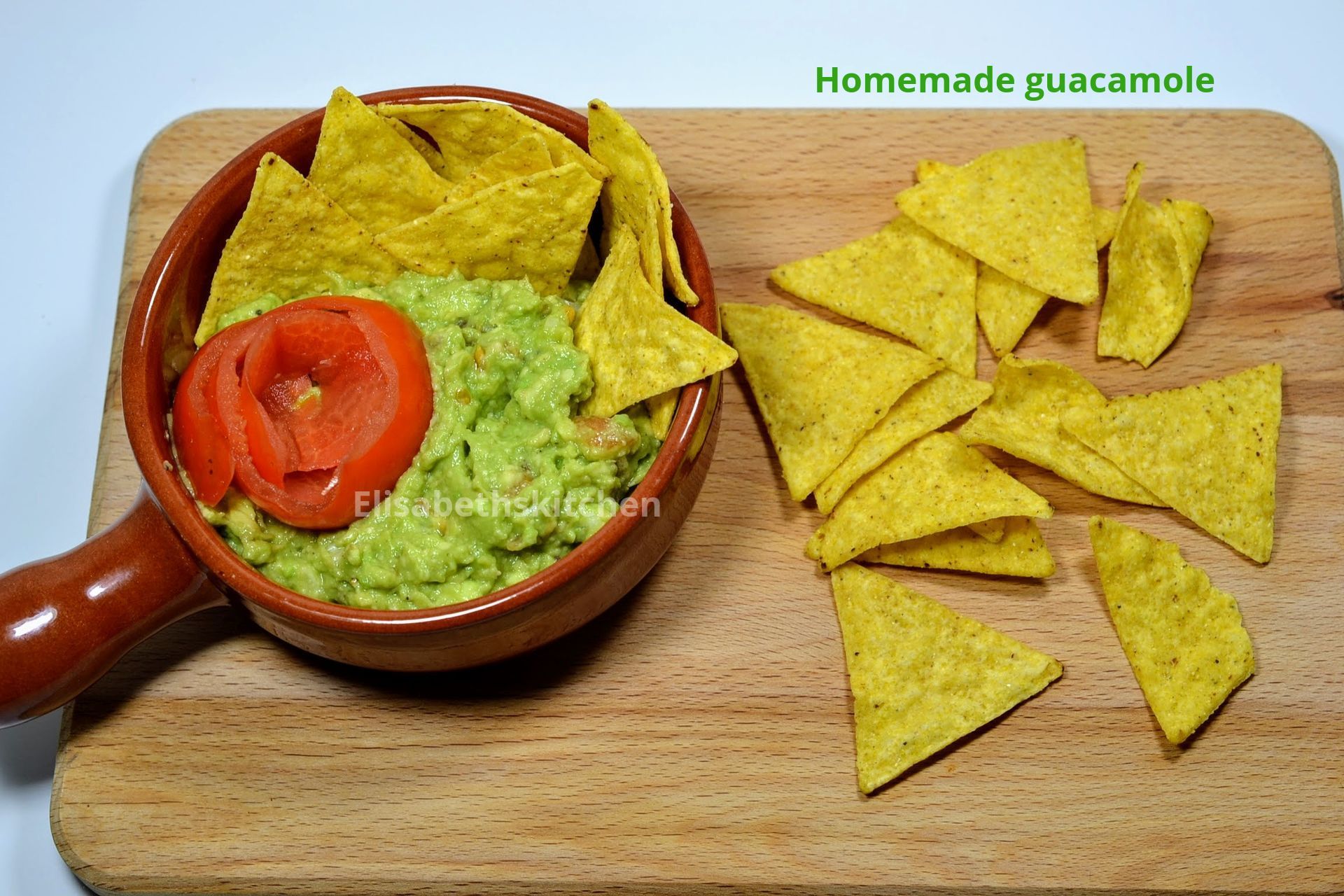 Scroll door voor het recept Guacamole in een kom met tortillachips op een houten plank. Een tomaatje als garnering is zichtbaar.
