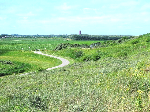 Een kronkelend pad door met gras begroeide duinen, met in de verte een groen veld en een rode toren onder een blauwe hemel.