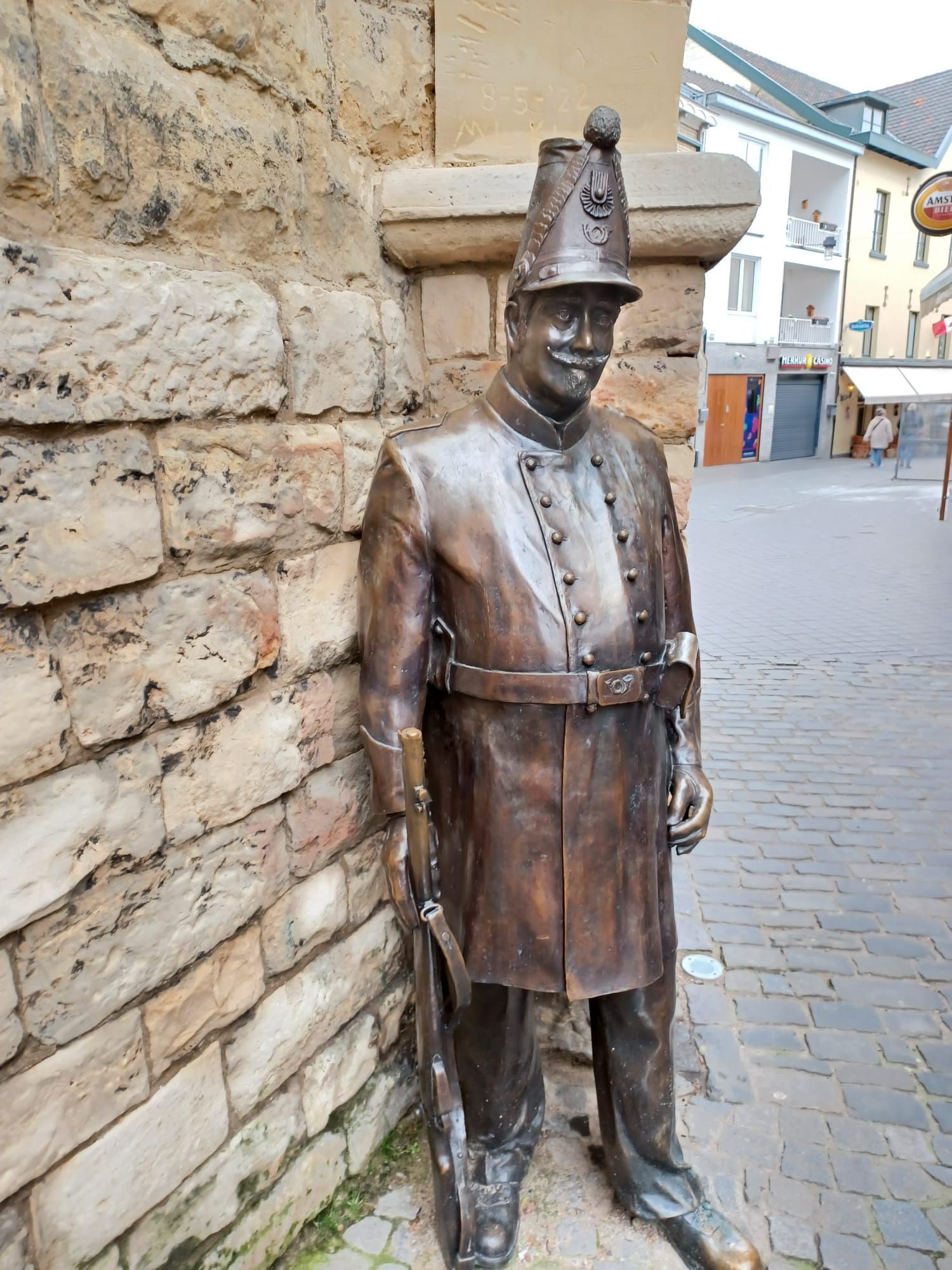 Bronzen beeld van een man in uniform die tegen een stenen muur leunt, in een straat met bakstenen bestrating.