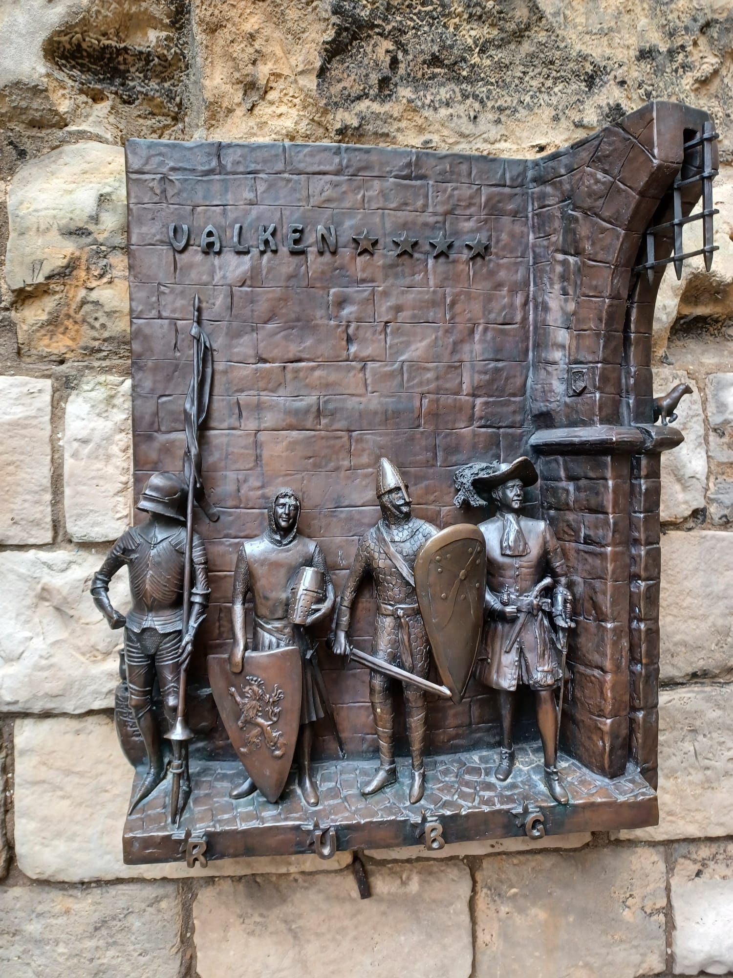 Bronzen plaquette met vier figuren: een soldaat in harnas, soldaten met schilden en zwaarden tegen een bakstenen muur.