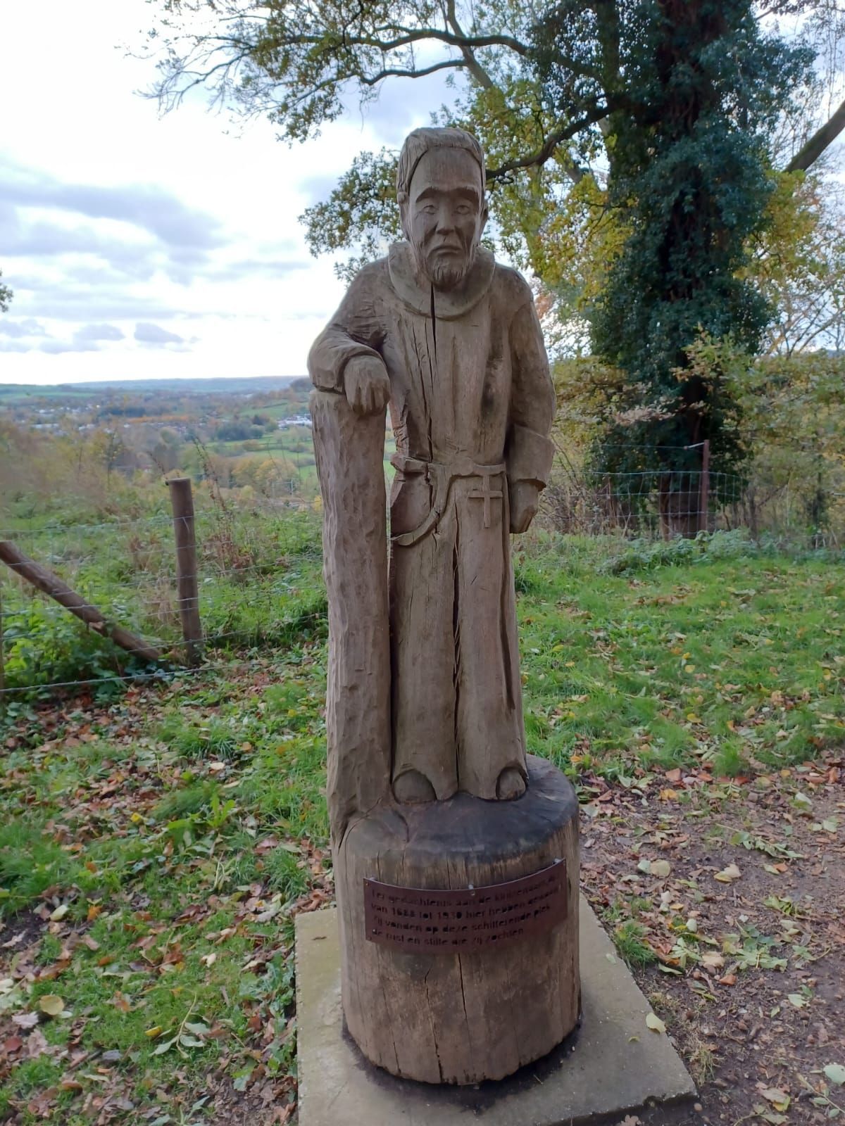 Houten beeld van een man in gewaad, leunend tegen een boomstam. Het bruine, verweerde houtsnijwerk staat op een stronk met een opschrift in de buitenlucht.