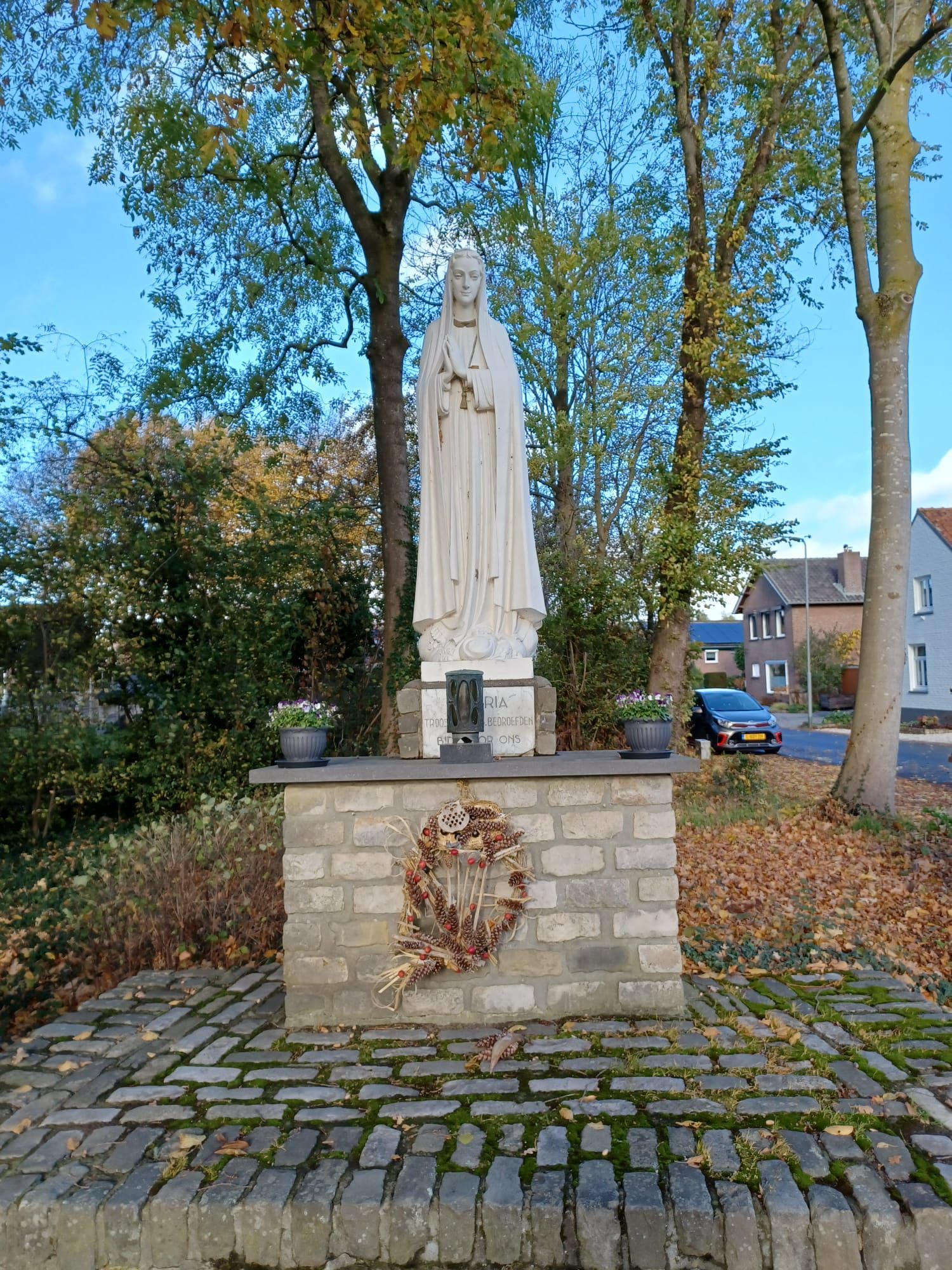 Standbeeld van een heilige op een bakstenen sokkel, met een krans en potplanten, in een buitenomgeving.